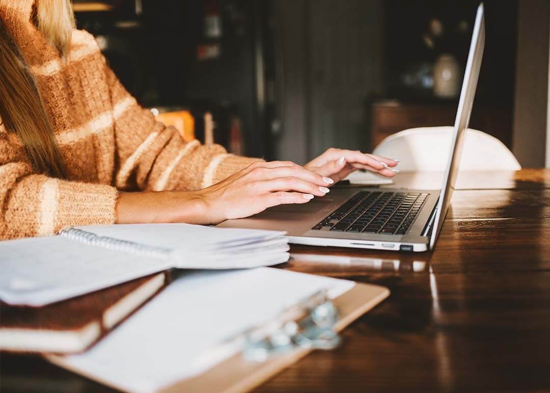 A woman wearing an orange jumper, working on a desk with open notebooks and a laptop, reviewing her finances.