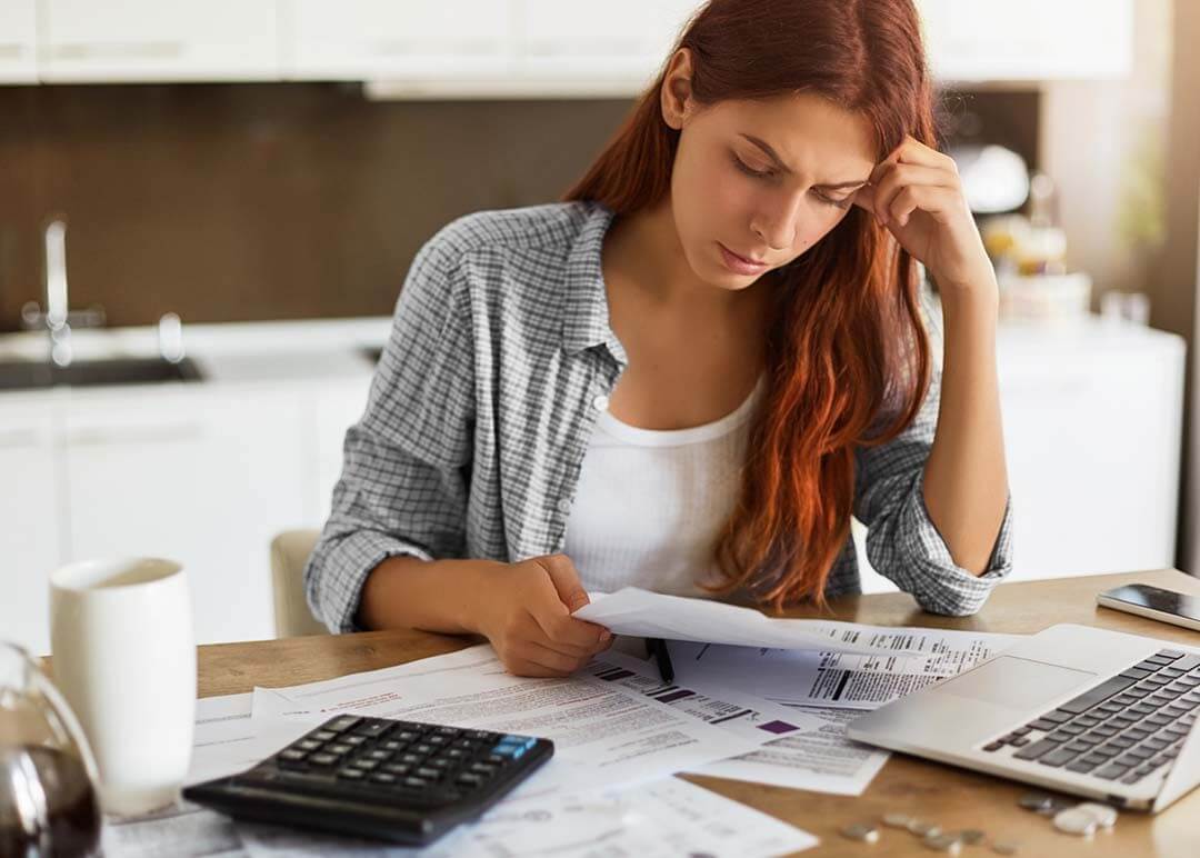 A young woman sits at her table, with a calculator, paperwork and laptop, worried about her finances. 